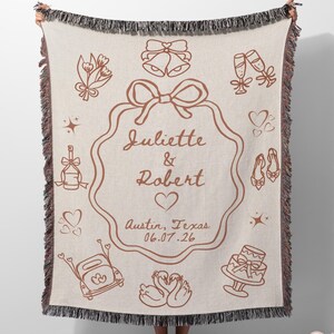 May include: A beige woven throw blanket with a brown wedding-themed design. The blanket features the names "Juliette & Robert", a heart, and the date 06.07.26. Decorative elements include bells, champagne glasses, and a cake.
