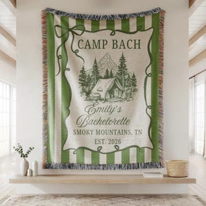 May include: A woven tapestry with green stripes and fringe. The center features a camping scene with a tent, trees, and mountains, along with the text "Camp Bach" and "Emily's Bachelorette, Smoky Mountains, TN, EST. 2026."