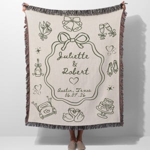 May include: A beige woven throw blanket with a dark green wedding-themed design. The design includes a central frame with the names "Juliette & Robert", a heart, and the date "06.07.26". The blanket has a fringed edge.