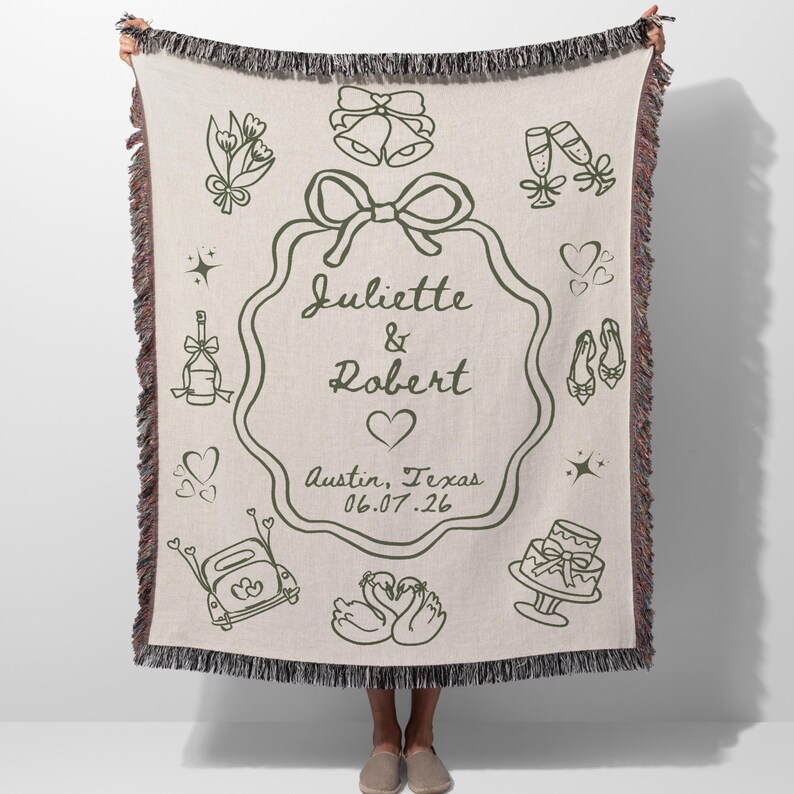 May include: A beige woven throw blanket with a dark green wedding-themed design. The design includes a central frame with the names "Juliette & Robert", a heart, and the date "06.07.26". The blanket has a fringed edge.