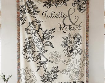 May include: Cream-colored woven throw blanket with black floral designs and fringe trim. The blanket features the names "Juliette & Robert" and the date "September 30, 2026". The floral pattern includes peonies and other flowers.