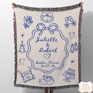 May include: Cream-coloured woven throw blanket with a blue wedding design. The blanket features illustrations of wedding bells, champagne flutes, hearts, and a cake. The names "Juliette & Robert" are displayed, along with the date 06.07.26.