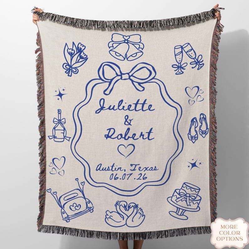 May include: Cream-coloured woven throw blanket with a blue wedding design. The blanket features illustrations of wedding bells, champagne flutes, hearts, and a cake. The names "Juliette & Robert" are displayed, along with the date 06.07.26.