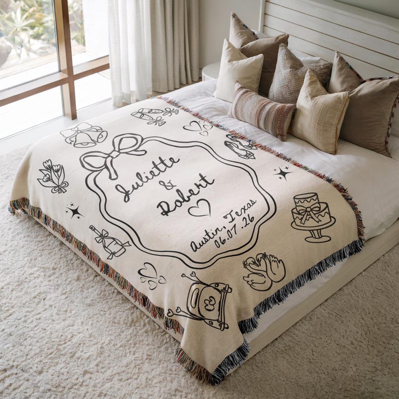 May include: Cream-coloured woven throw blanket with a black outline design. The blanket features a central framed design with the names "Juliette & Robert", a heart, and the date "06.07.26" and location "Austin, Texas". The blanket has a fringed edge.