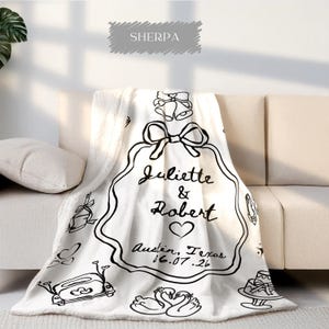 May include: A white sherpa blanket with black line art. The blanket features a large decorative frame with the names "Juliette & Robert", a heart, and the date "16.07.26" and location "Austin, Texas".