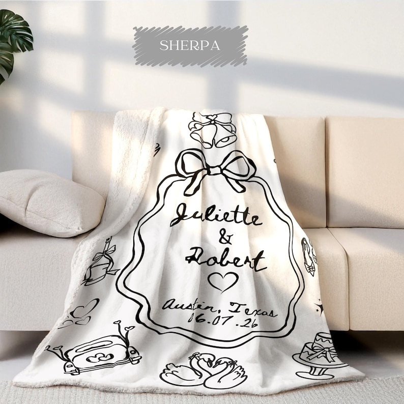 May include: A white sherpa blanket with black line art. The blanket features a large decorative frame with the names "Juliette & Robert", a heart, and the date "16.07.26" and location "Austin, Texas".