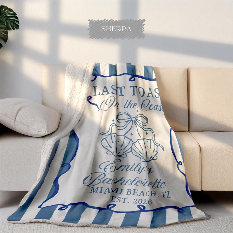 May include: A white Sherpa blanket with blue stripes and text that reads "LAST TOAS On the Coast Emily's Bachelorette MIAMI BEACH, FL EST. 2026". The blanket features seashell graphics and a bow design.