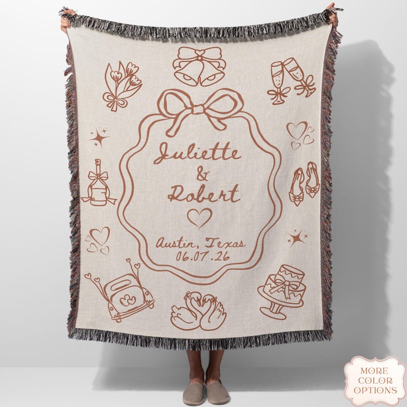 May include: A beige woven throw blanket with a brown wedding-themed design. The blanket features the names "Juliette & Robert", a heart, and the date 06.07.26. Decorative elements include bells, champagne glasses, and a cake.