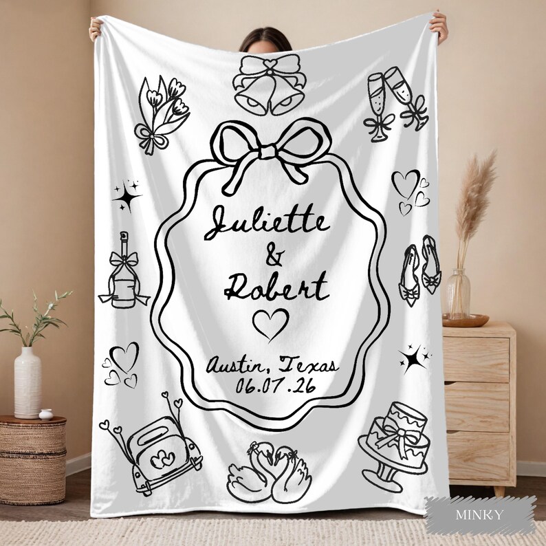 May include: White blanket with black line art illustrations of wedding-themed icons, including bells, champagne flutes, and a cake. The blanket features the names "Juliette & Robert" and the date "06.07.26" in Austin, Texas.
