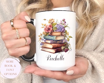 May include: White ceramic mug with a black handle and rim, featuring a watercolor illustration of a stack of books adorned with flowers. The name "Rachelle" is written in elegant script below the books. "More book & font options" is written on the bottom left.