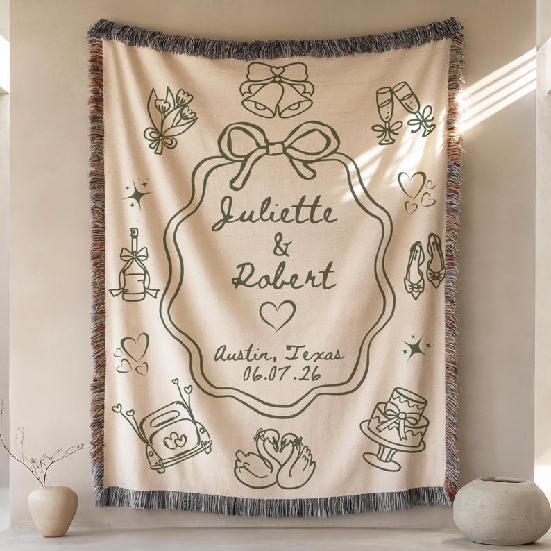 May include: Cream-coloured woven throw blanket with a dark green decorative design. The design includes the names "Juliette & Robert", a heart, and the date 06.07.26, along with wedding-themed illustrations such as bells and a cake.