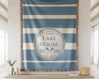 May include: Blue and cream striped throw blanket with fringed edges. The center features a circular emblem with the text "Lake House" and "The Thompson Family" around the edge, with crossed oars and waves. The blanket is dated "Est. 2026".