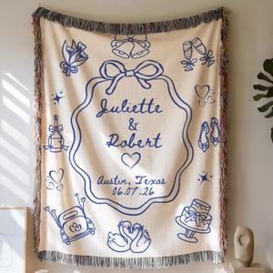 May include: Cream-coloured woven throw blanket with blue illustrations and fringe. The blanket features a central design with the names "Juliette & Robert", a heart, and the date 06.07.26. Additional illustrations include wedding-themed icons.