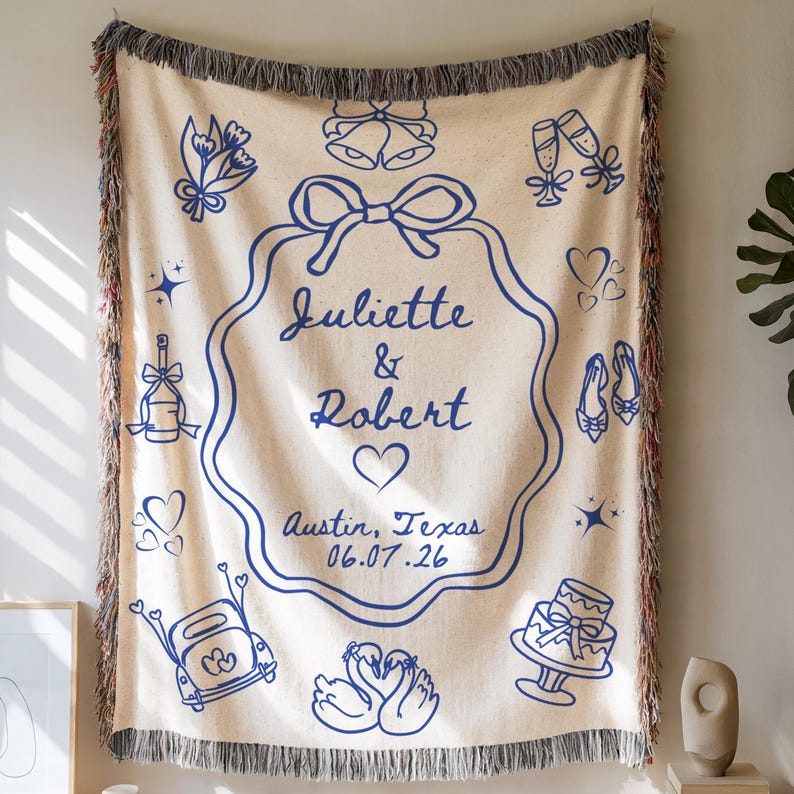 May include: Cream-coloured woven throw blanket with blue illustrations and fringe. The blanket features a central design with the names "Juliette & Robert", a heart, and the date 06.07.26. Additional illustrations include wedding-themed icons.