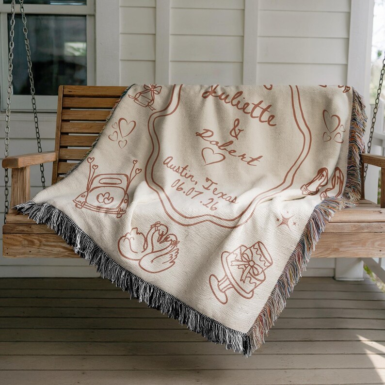 May include: A beige woven blanket with brown illustrations and text, draped over a wooden swing. The blanket features the names "Juliette & Robert", a heart, and the date "06.07.26" with the location "Austin, Texas". The blanket has a fringed edge.