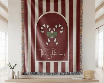 May include: A red and white striped throw blanket with a festive candy cane design. The design features two crossed candy canes with a green bow, and the text "The Johnsons" in a script font. The blanket has a fringed edge.