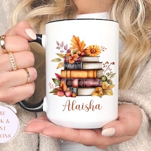 May include: White ceramic mug with a black handle and rim, featuring a watercolor design of stacked books, autumn leaves, and a sunflower. The name "Alaisha" is printed on the mug. "More Book & Font Options" is also printed on the mug.