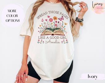 May include: Ivory-colored t-shirt with a floral and butterfly design. The graphic includes an open book with flowers and the text "SPREAD THOSE PAGES LIKE A GOOD GIRL Amelia". The shirt has short sleeves.