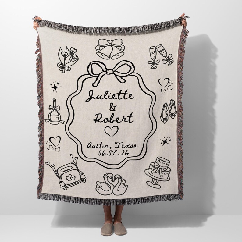 May include: Cream-coloured woven throw blanket with black line art illustrations of wedding-themed icons. The blanket features the names "Juliette & Robert", a heart, and the date 06.07.26, along with the location "Austin, Texas".
