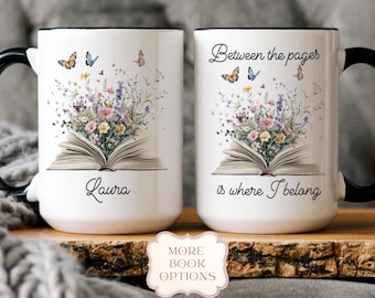May include: Two white ceramic mugs with black handles and rims. Each mug features an open book design with colorful flowers and butterflies. One mug says "Laura", the other says "Between the pages is where I belong."