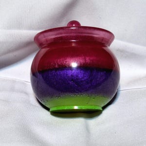 May include: A small, round, three-layered resin jar with a lid. The jar is green, purple, and pink.