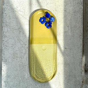 May include: A yellow, pill-shaped ceramic tray with a textured surface. The top of the tray holds three blue flower-shaped objects. The tray is resting on a textured, light-colored surface.