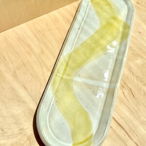 May include: An elongated, oval-shaped ceramic tray with a glossy off-white glaze and a speckled finish. A single, wide, translucent yellow brushstroke pattern curves diagonally across the center of the tray from the top edge to the bottom.
