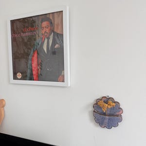 May include: A framed album cover featuring "el chico chico hamilton" in red text. The image shows a man in a suit. A decorative, blue and gold ceramic wall hanging is also visible.