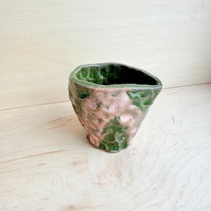 May include: A small, hand-formed ceramic cup with an irregular, organic shape. The exterior features a glossy, textured glaze in deep forest green with patches of soft, muted pink. The rim is uneven, and the interior is finished in a dark green glaze.