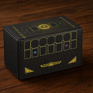 May include: A rectangular black storage box featuring gold-colored graphic prints. The top displays a central eye motif with radiating lines. The front side shows a grid of rectangles, a winged scarab beetle, and two small colored gem icons.
