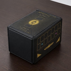 May include: A rectangular black deck box featuring gold foil accents. The top lid displays a central eye symbol surrounded by radiating lines and hieroglyphic-style text. The side panel shows a grid pattern with a golden scarab beetle icon.