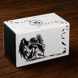 May include: A rectangular storage box with a white exterior and black side panels. The top surface features a black Japanese character inside a sunburst design. The front panel displays a detailed black ink illustration of a winged creature.