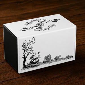May include: A rectangular deck box with a white exterior and black side panels. The top features a circular emblem surrounded by floral patterns. The front displays a black ink-style illustration of a frog near a tree, a creature, and foliage.