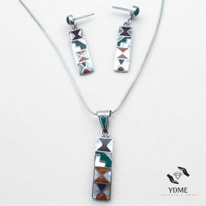Op de afbeelding: Een zilveren ketting- en oorbellenset. De ketting heeft een hanger met een rechthoekig ontwerp, versierd met kleurrijke geometrische vormen. De bijpassende oorbellen hebben een vergelijkbaar ontwerp. De sieradenset wordt op een witte achtergrond gepresenteerd.