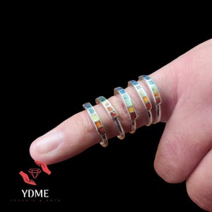 Puede incluir: Cinco anillos de plata con incrustaciones de esmalte de colores. Los anillos están apilados en un dedo, mostrando las diferentes combinaciones de colores.