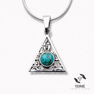 Puede incluir: Un colgante triangular de plata con una piedra turquesa redonda en el centro. El marco de metal tiene detalles recortados que representan pequeñas figuras de animales. El colgante cuelga de una fina cadena de serpiente, con el texto YDME JOYERIA & ARTE.