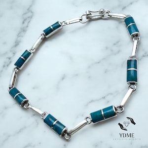May include: Silver bracelet with rectangular turquoise stones and silver detailing. The bracelet is made of linked sections and has a clasp. The jewellery is shown on a marble surface.