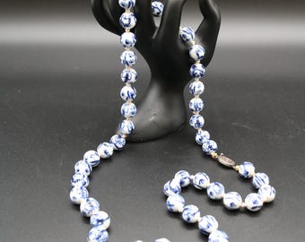 May include: A necklace with round, white beads featuring a blue floral design. The beads are strung together with a light-colored string and are displayed on a black hand-shaped stand against a black background.
