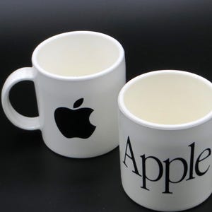 Op de afbeelding: Twee witte keramische mokken met zwarte bedrukking. Eén mok heeft het zwarte Apple-logo op de voorzijde, terwijl de andere mok het woord Apple in een zwart schreeflettertype toont. Beide mokken hebben een gladde afwerking en een zijhandvat.