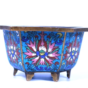 Puede incluir: Un cuenco decorativo hexagonal azul con diseños florales en rosa, rojo y blanco. El cuenco tiene un borde dorado y pequeños pies. El patrón floral está centrado en cada lado del cuenco.