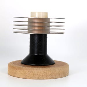 May include: A desk accessory featuring a circular light-colored wooden base, a black cylindrical central post, and a top section composed of five stacked, thin, metallic silver discs surrounding a small, pale, cylindrical center piece.