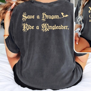 May include: A dark gray t-shirt with the text "Save a Dragon. Ride a Mingleader." printed in gold on the back. The front of the shirt has the text "Xaddy (zædi) noun xaden riorson" printed in gold.