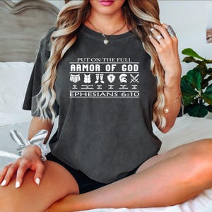 May include: A dark gray t-shirt with a white graphic that says "Put on the full Armor of God" with a list of different armor pieces and the scripture reference "Ephesians 6:10".