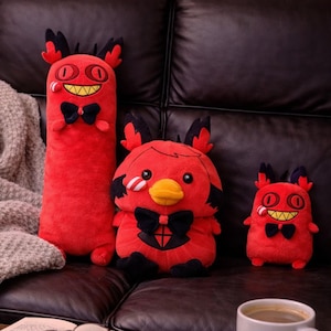 May include: Three red plush toys shaped like cartoon demons with black details and bow ties. One is tall and cylindrical, another is round with a yellow beak, and the third is small. They are on a brown leather sofa.