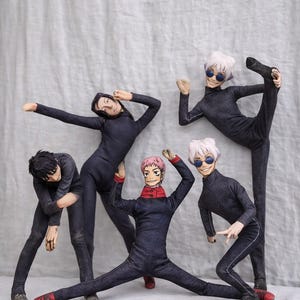 May include: Five dolls in dark blue jumpsuits are posed in various athletic positions against a grey backdrop. The dolls have different hairstyles and facial features, some wearing sunglasses and red accents on their shoes and collars.