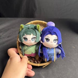 Könnte beinhalten: Zwei Plüschpuppen mit Anime-Stil-Merkmalen, eine in Grün und die andere in Blau, sitzen in einem kleinen Weidenkorb mit Kaffeebohnen. Die Puppen haben große Augen und sind in traditioneller Kleidung gekleidet.