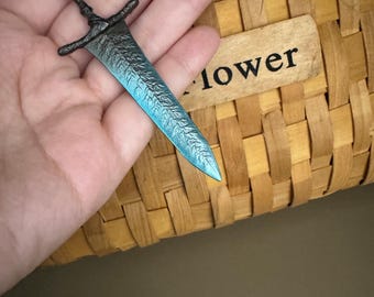 May include: A blue and silver sword keychain with a detailed design. The keychain is hanging from a hand.