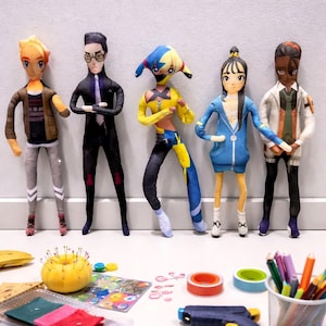 May include: Five fabric dolls with distinctive outfits and hairstyles stand against a grey wall. The dolls have painted faces. Craft supplies, including pins, tape, and coloured pencils, are scattered on the white surface in front of the dolls.