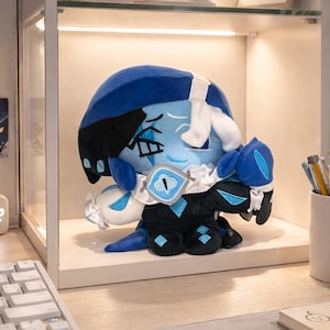 May include: A plush doll with a blue and black color scheme, featuring a stylized design. The doll has a blue head, black accents, and white details. It is displayed inside a glass-fronted cabinet, suggesting a collectible or decorative item.