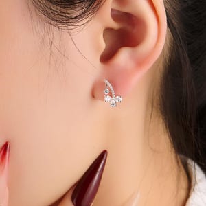 May include: A close-up of a silver earring with a butterfly design. The earring features small, clear gemstones arranged in a curved shape, with a larger gemstone at the center of the butterfly. The earring is worn on a person's ear.